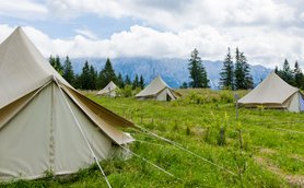 Eine Gruppe von Zelten steht auf einer grünen Wiese, umgeben von hohen Bäumen und majestätischen Bergen im Hintergrund.