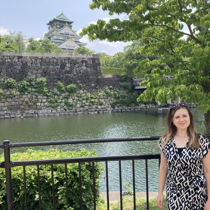 Eine Frau in einem gemusterten Kleid steht am Ufer eines ruhigen Gewässers, im Hintergrund ist eine beeindruckende Burg zu sehen.