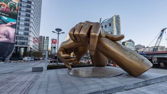 Eine große, goldene Statue einer Hand, die ein Schild mit der Aufschrift 'Gangnam Style' hält, steht in einer urbanen Umgebung.