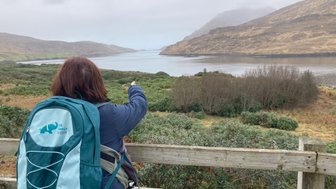 Eine Person mit einem Rucksack zeigt auf die ruhige Killary Fjord-Landschaft, umgeben von sanften Hügeln und bewaldeten Ufern.