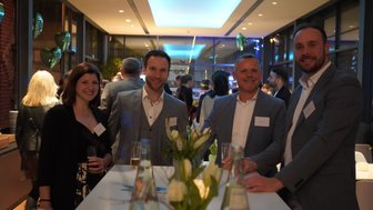 Eine Gruppe von vier Personen lächelt an einem eleganten Tisch in einem modernen Veranstaltungsraum mit Pflanzen und Getränken.