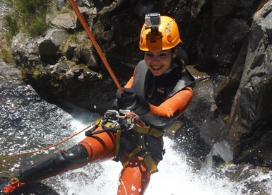 Eine Person in einem orangefarbenen Neoprenanzug seilt sich an einem Wasserfall ab, umgeben von spritzendem Wasser.