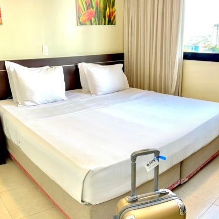 Ein helles Hotelzimmer mit einem großen Bett, zwei Kissen und einem Koffer neben dem Bett auf dem Boden.