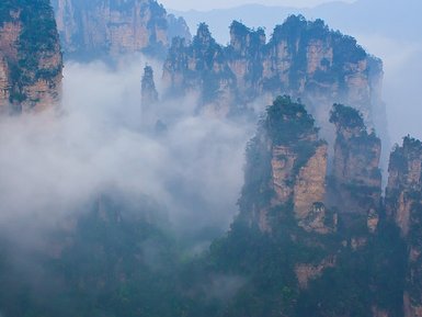 Majestätische, nebelverhangene Felsen ragen aus einem dichten Wald, umgeben von mystischer Atmosphäre und sanften Farben.