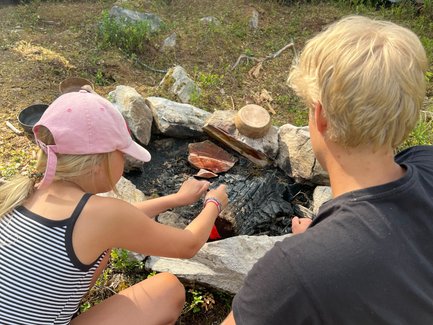 Ein Mädchen in einem gestreiften Shirt grillt über einem Lagerfeuer, während ein Junge daneben zuschaut und sich konzentriert.