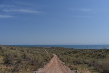 Ein schmaler, staubiger Weg schlängelt sich durch eine grüne Landschaft mit Blick auf einen ruhigen See und blauen Himmel.