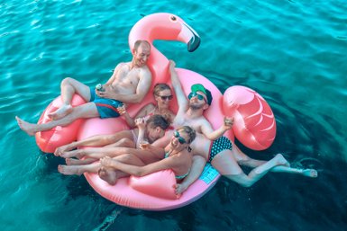 Fünf Freunde entspannen auf einem großen, pinken Flamingo-Schwimmring im klaren Wasser, lachen und genießen Getränke.