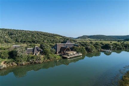 Ein malerisches Flussufer mit einer Lodge, umgeben von üppigem Grün und sanften Hügeln unter einem klaren blauen Himmel.