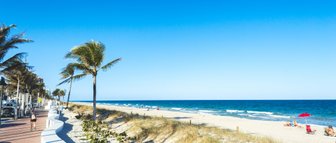 Ein sonniger Strand in Fort Lauderdale mit Palmen, einem klaren blauen Himmel und Menschen, die sich am Wasser entspannen.