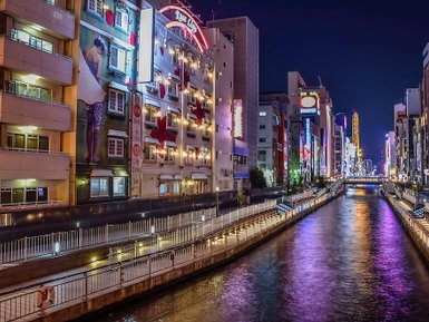 Die nächtliche Skyline von Osaka spiegelt sich im ruhigen Wasser eines Kanals, umgeben von bunten Neonlichtern und Gebäuden.
