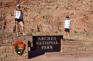 Zwei Kinder stehen auf einem Steinblock vor dem Eingang zum Arches Nationalpark, umgeben von beeindruckenden roten Felsen.