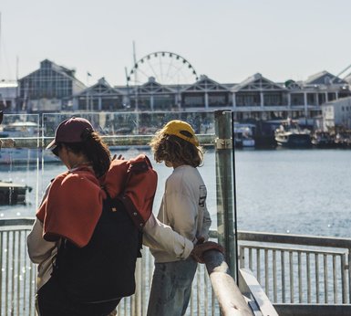 Zwei Kinder stehen am Wasser, eines trägt einen Rucksack und einen roten Pullover, während das andere den Blick über das Wasser schweifen lässt.