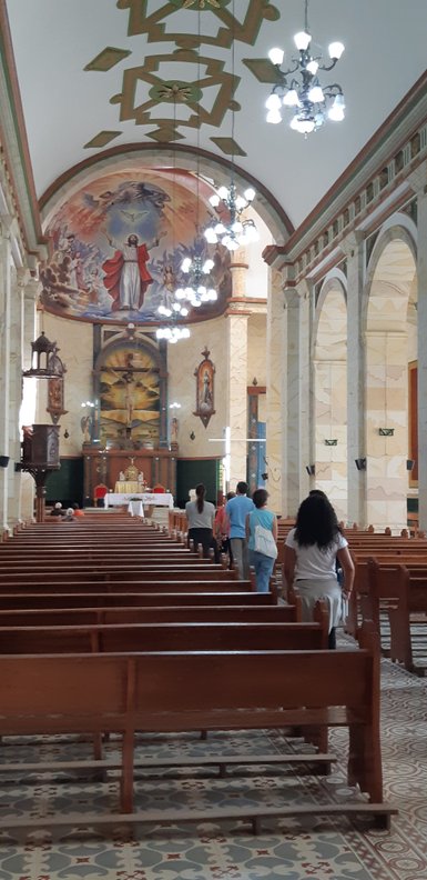 Die Innenansicht einer Kirche mit bunten Wandmalereien und Holzbankreihen, in denen Menschen beten und sich versammeln.