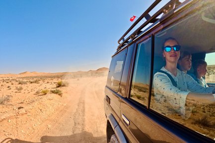 Eine Frau mit Sonnenbrille fährt in einem Geländewagen durch die Wüste, während zwei Kinder auf dem Beifahrersitz sitzen.