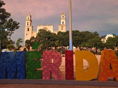 Bunte Buchstaben bilden das Wort 'Mérida' vor einer beeindruckenden historischen Architektur unter einem farbenfrohen Himmel.