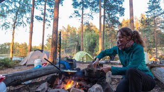 Eine Frau kocht am Lagerfeuer in einem Waldcamp, umgeben von Zelten und Bäumen, während die Sonne untergeht.