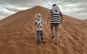 Ein Erwachsener und ein Kind gehen Hand in Hand einen hohen Sanddünenhang hinauf, umgeben von einer weiten Wüstenlandschaft.