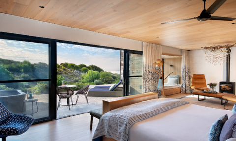 Ein modernes Schlafzimmer mit Holzdecke, großen Fenstern und Blick auf eine grüne Landschaft und den Ozean im Hintergrund.