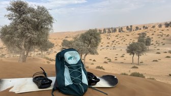 Ein Rucksack und ein Snowboard liegen auf einer Sanddüne, umgeben von vereinzelten Bäumen in einer Wüstenlandschaft.