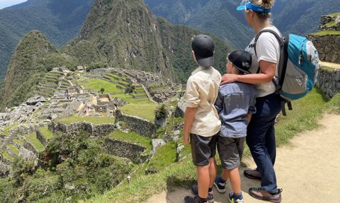 Eine Frau steht mit zwei Kindern an einem Aussichtspunkt und bewundert die Ruinen von Machu Picchu in der Ferne.