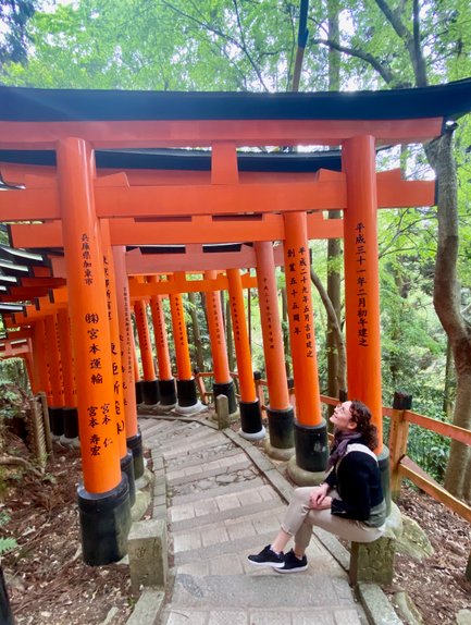 Eine Person sitzt auf einer Steinbank und blickt lächelnd zu den leuchtend orangefarbenen Torii-Toren auf einem schmalen Weg.