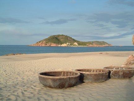 Drei runde Fischerboote aus Korbgeflecht stehen am sandigen Strand, während eine grüne Insel im Hintergrund sichtbar ist.