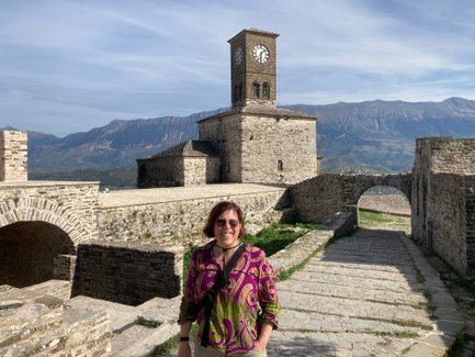 Eine Frau mit braunen Haaren steht lächelnd vor einer alten Steinburg mit einem Uhrturm und Bergen im Hintergrund.