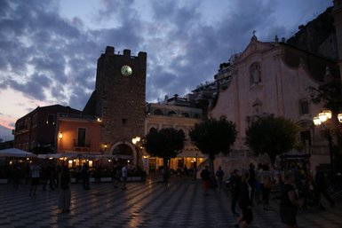 Der Platz in Taormina ist bei Dämmerung beleuchtet, mit einem historischen Turm und einer Kirche im Hintergrund.