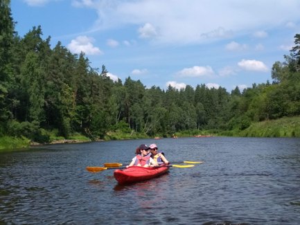 Zwei Personen im Kajak paddeln auf einem ruhigen Fluss, umgeben von üppigem Grün und blauen Himmel.