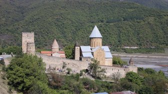 Eine beeindruckende Festung mit mehreren Türmen und einer Kirche, umgeben von üppigen grünen Bergen und einem klaren blauen Himmel.