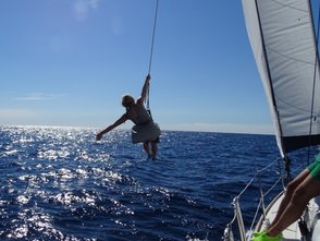 Eine Person schwingt fröhlich über dem glitzernden Wasser, während sie an einem Seil von einem Segelboot hängt.