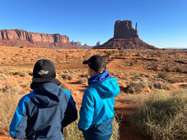 Zwei Jungen stehen in der Wüste und betrachten die beeindruckenden Felsformationen von Monument Valley unter klarem Himmel.