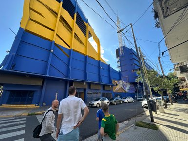 Drei Personen stehen auf der Straße und betrachten das beeindruckende, blau-gelbe Stadiongebäude in Buenos Aires.