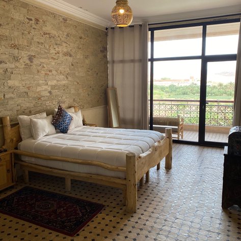 Geräumiges Zimmer mit einem Holzbett, Kunstwerken auf dem Bett und einem Blick auf die grüne Landschaft durch große Fenster.