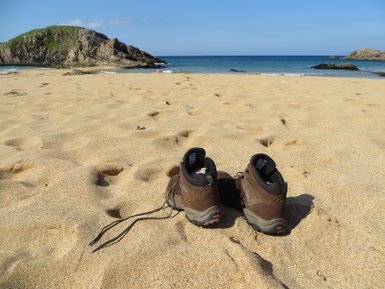 Am Strand stehen ein Paar Wanderschuhe im Sand, während sanfte Wellen an die Küste schlagen und eine grüne Hügelkuppe im Hintergrund sichtbar ist.