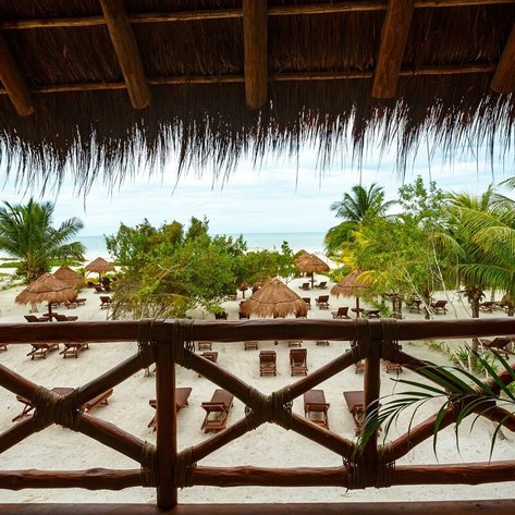 Blick von einer Holzveranda auf einen ruhigen Strand mit Liegen und Palmen, umgeben von schattigen Hütten.