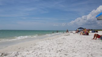 Ein sonniger Strand mit feinem, weißem Sand, wo Menschen im Wasser spielen und sich entspannen.