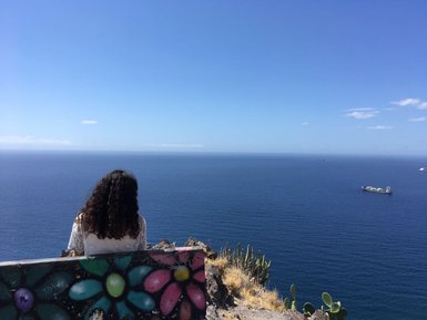 Eine Person mit lockigem Haar sitzt auf einem Felsen und blickt auf das weite, ruhige Meer unter einem strahlend blauen Himmel.