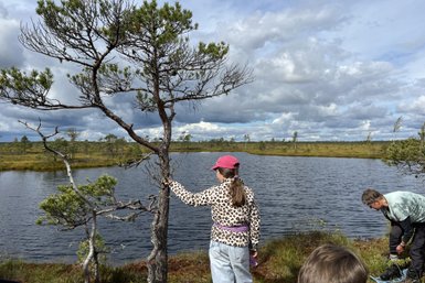 Ein Mädchen mit einem pinken Hut steht an einem Gewässer und berührt einen Baum, während Wolken den Himmel bedecken.