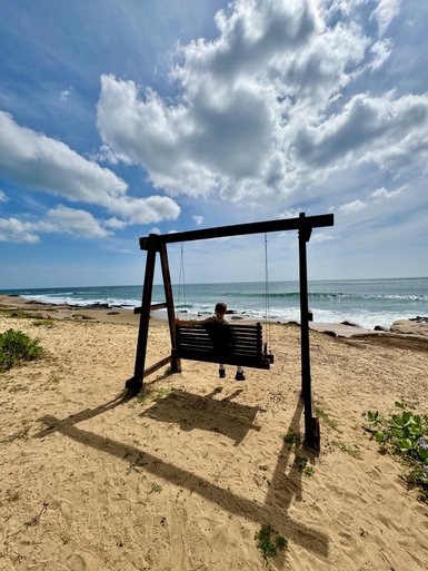 Eine Person sitzt auf einer Schaukel am Strand, umgeben von sanften Wellen und einem klaren, blauen Himmel.