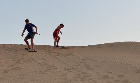 Zwei junge Männer gleiten auf Sandboards die sanften Dünen einer Wüste hinunter, umgeben von goldenem Sand und klarem Himmel.