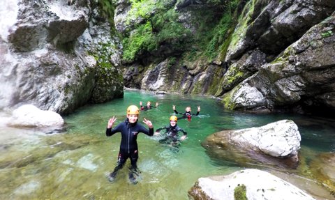 Eine Gruppe von Menschen in Neoprenanzügen genießt das Wasser eines klaren Flusses, umgeben von hohen Felsen und Bäumen.