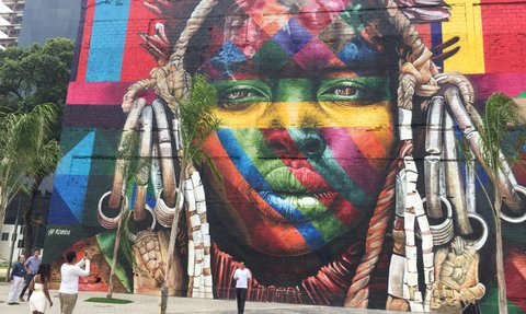 Bunter Wandmural zeigt das Gesicht einer Frau mit leuchtenden Farben und traditionellen Schmuckstücken, umgeben von Palmzweigen.