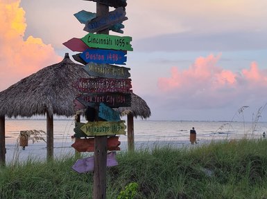 Ein bunter Wegweiser mit Entfernungen zu verschiedenen Städten steht am Strand, während die Sonne untergeht.