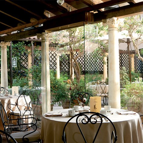 Ein elegantes Restaurant mit runden Tischen, umgeben von Pflanzen und einem Glasfenster, das den Garten zeigt.