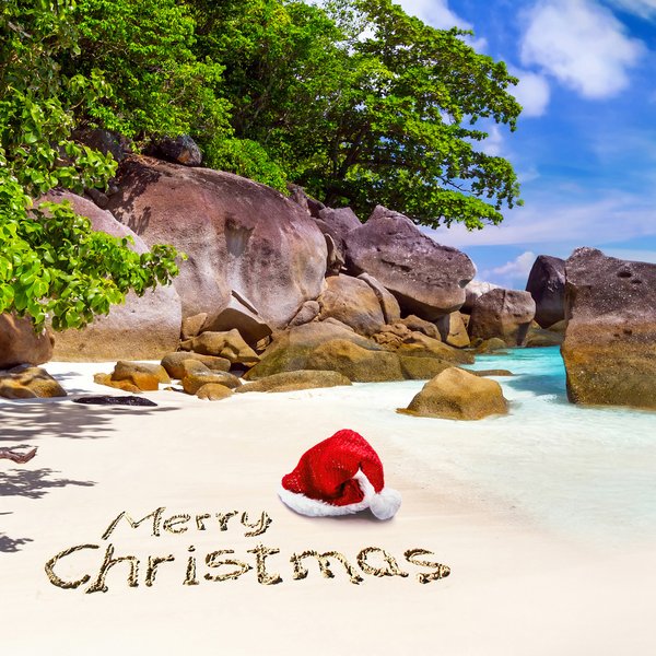 Ein roter Weihnachtsmann-Hut liegt am Strand, während 'Frohe Weihnachten' im Sand geschrieben steht, umgeben von Felsen und klarem Wasser.