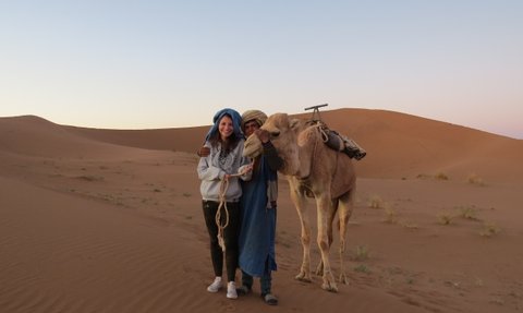 Teenagerin steht gemeinsam mit einem Beduinen neben einem Kamel in der Wüste – Familienurlaub in Marokko