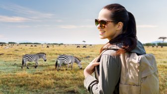 Eine lächelnde Frau mit Sonnenbrille steht in einer weiten Savanne, während Zebras im Hintergrund grasen.