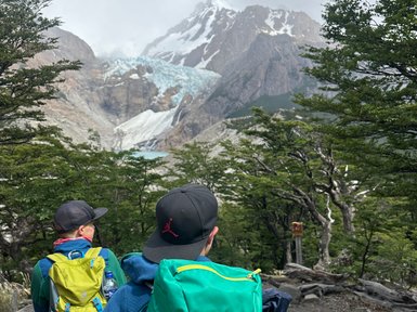 Zwei Wanderer mit Rucksäcken stehen auf einem Pfad, umgeben von Bäumen und Blick auf schneebedeckte Berge im Hintergrund.