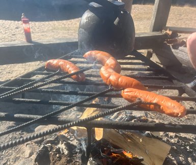 Über dem offenen Feuer brutzeln mehrere Würstchen auf einem Grillrost, während Rauch in die Luft steigt.